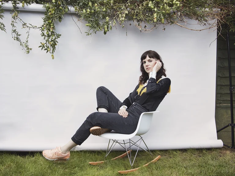 Eine Frau (Anna Meredith) sitzt lässig auf einem Stuhl vor einem neutralen Hintergrund im Freien, umgeben von Gräsern und grünen Zweigen.