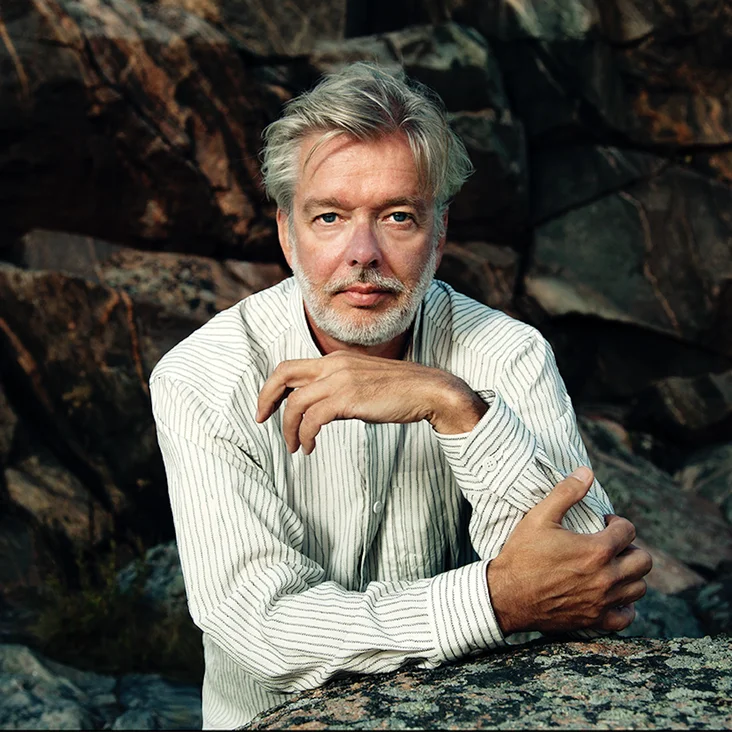 Jukka-Pekka Saraste: Ein Mann mit grauem Haar und Bart in einem weißen Hemd sitzt in einer felsigen Landschaft.