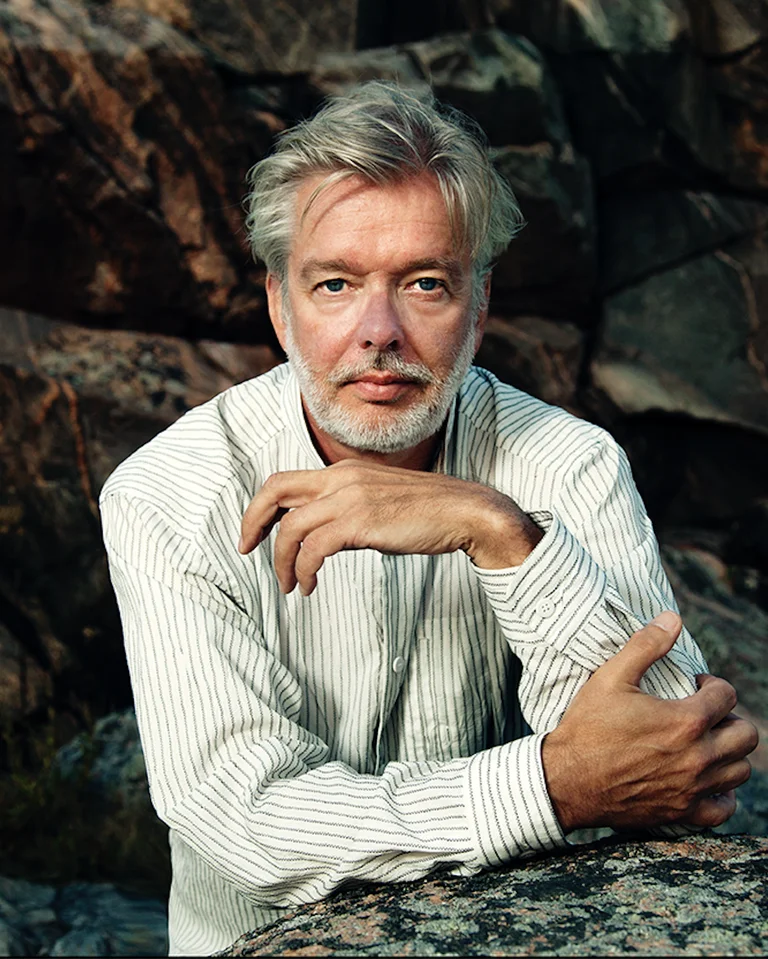 Jukka-Pekka Saraste: An man with gray hair and a beard, wearing a white shirt, sits in a rocky landscape.