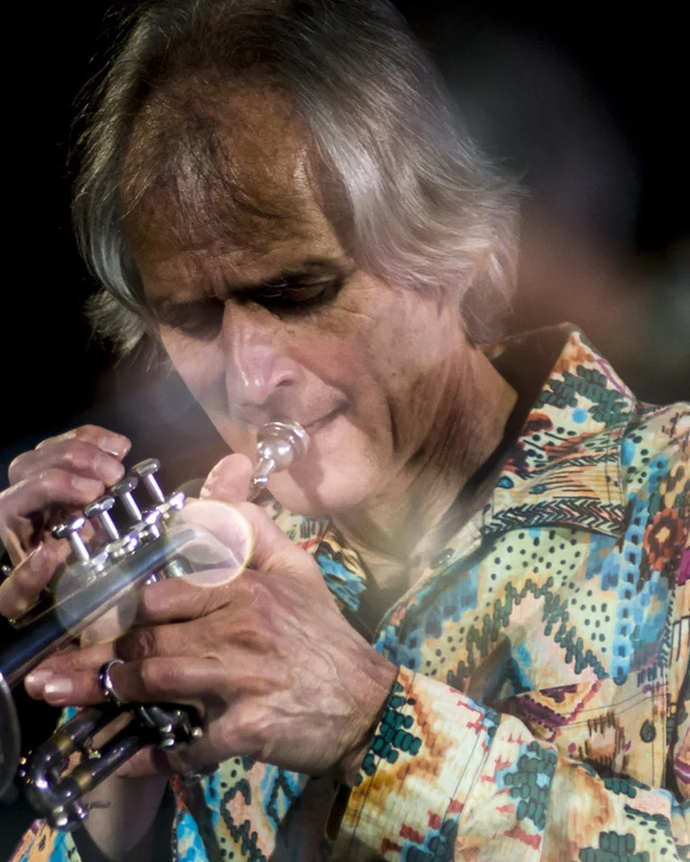 Ein Musiker (Markus Stockhausen) in einem bunten Hemd spielt leidenschaftlich Trompete vor einem dunklen Hintergrund, konzentriert auf seine Musik.