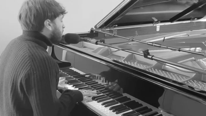 Samora Pinderhughes, captured from a rear-side angle while playing a grand piano. The still image from the video is in black and white.