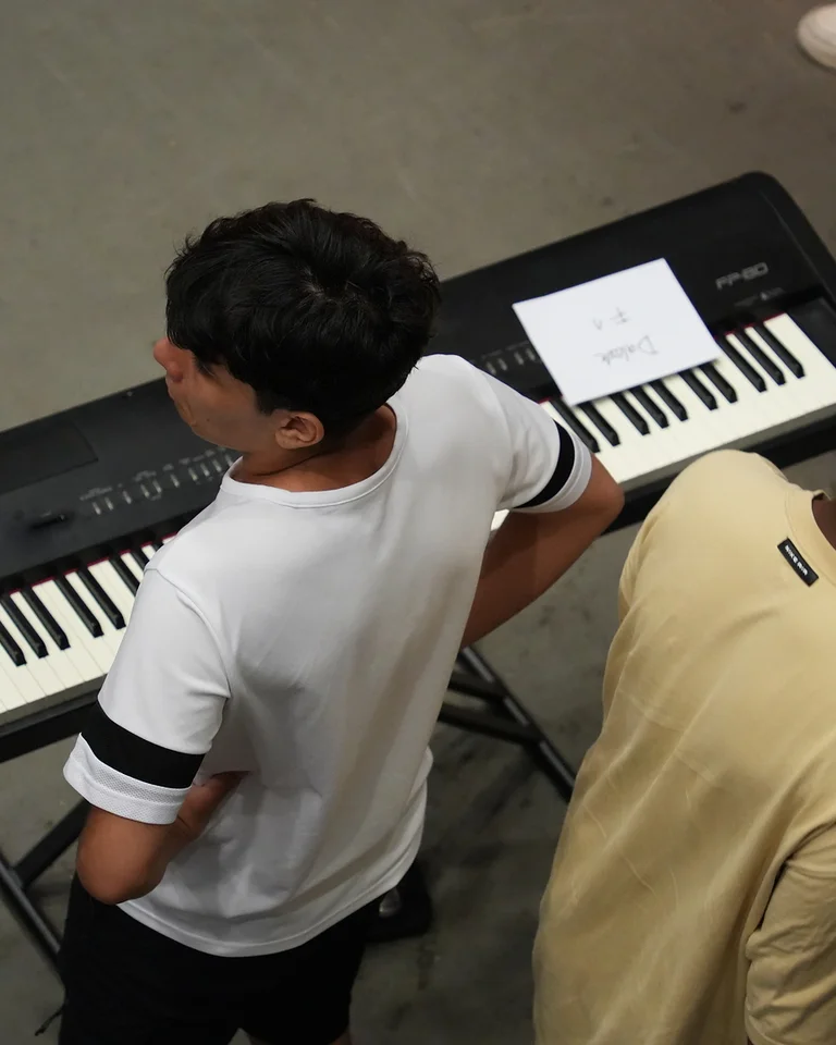 Two young people stand by an electronic keyboard with sheet music. They appear focused and ready to play.
