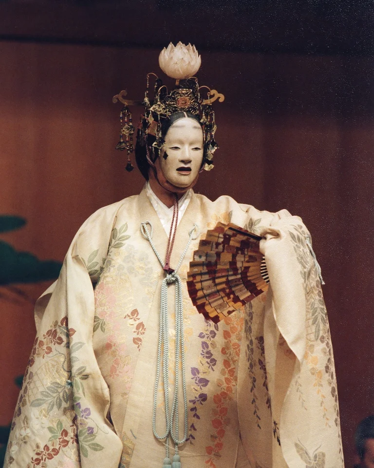 Ein traditioneller Noh-Schauspieler in aufwendigem Kostüm (Hagoromo) und Maske, hält einen Fächer. Der Hintergrund zeigt eine schlichte Bühne.
