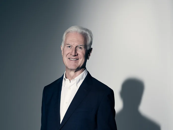 Ein lächelnder Mann in einem dunklen Anzug steht vor einem schattigen Hintergrund. Er hat graues Haar.