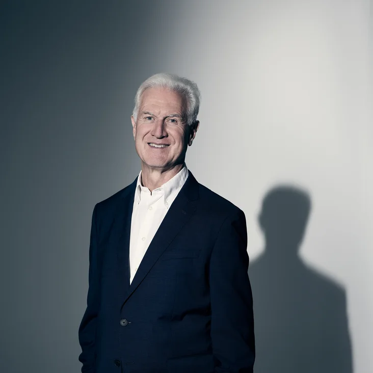 Ein lächelnder Mann in einem dunklen Anzug steht vor einem schattigen Hintergrund. Er hat graues Haar.