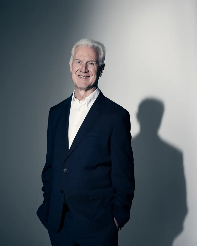 Ein lächelnder Mann in einem dunklen Anzug steht vor einem schattigen Hintergrund. Er hat graues Haar.