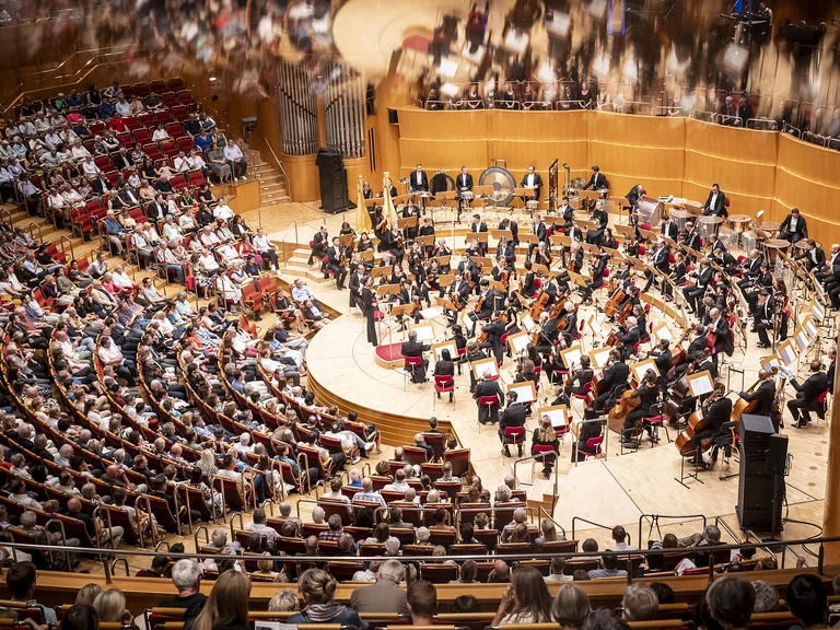 Ein vollbesetzter Konzertsaal mit einem Orchester (Gürzenich-Orchester Köln) auf der Bühne, das in einem großen Auditorium spielt. Zuschauende sitzen erwartungsvoll.