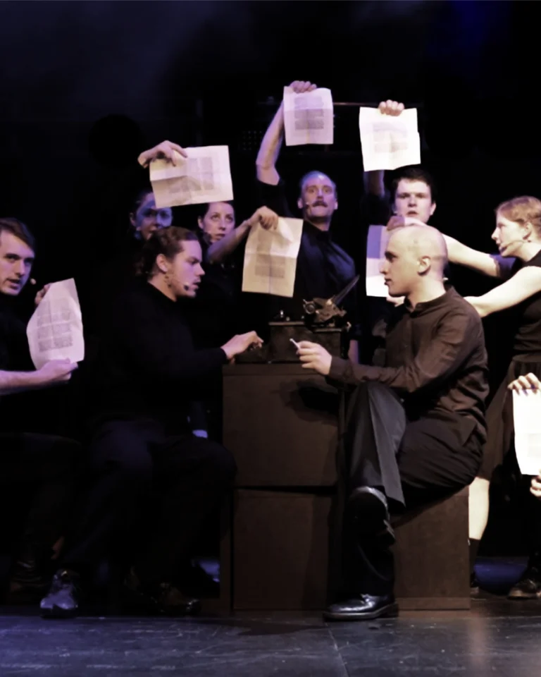 Eine Aufführung auf einer Bühne, bei der Personen in schwarzer Kleidung Papiere hochhalten. Die Szene ist dramatisch beleuchtet.