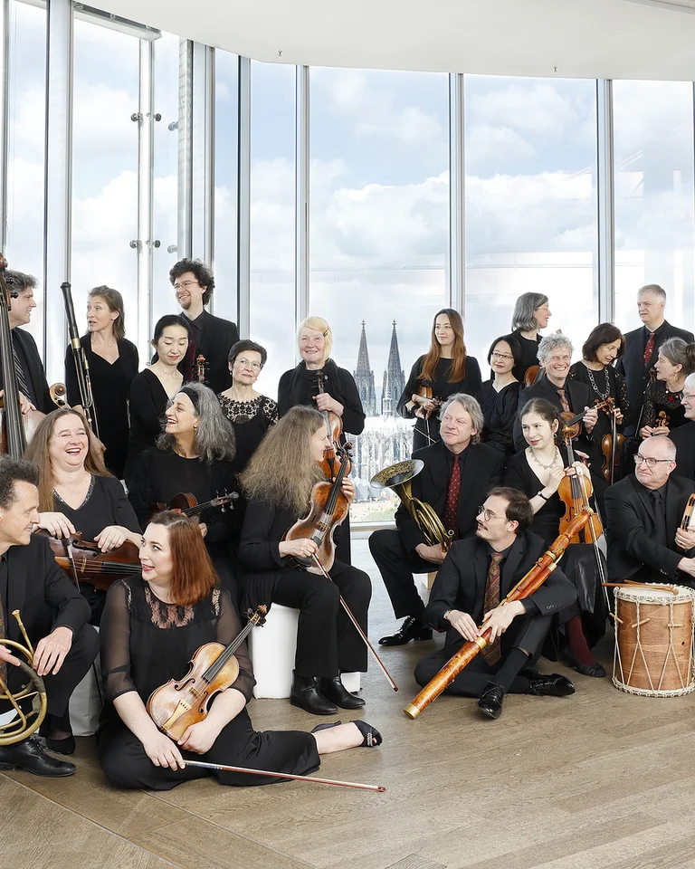 Ein Ensemble von Musikern (Concerto Köln) in einem modernen Raum mit Glaswänden. Sie halten verschiedene Musikinstrumente und tragen elegante schwarze Kleidung.