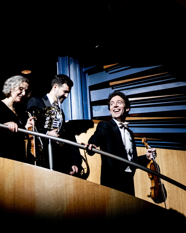 Vier festlich gekleidete Musiker steigen eine Treppe in einem Konzertsaal hinab, strahlend und mit Instrumenten in der Hand.