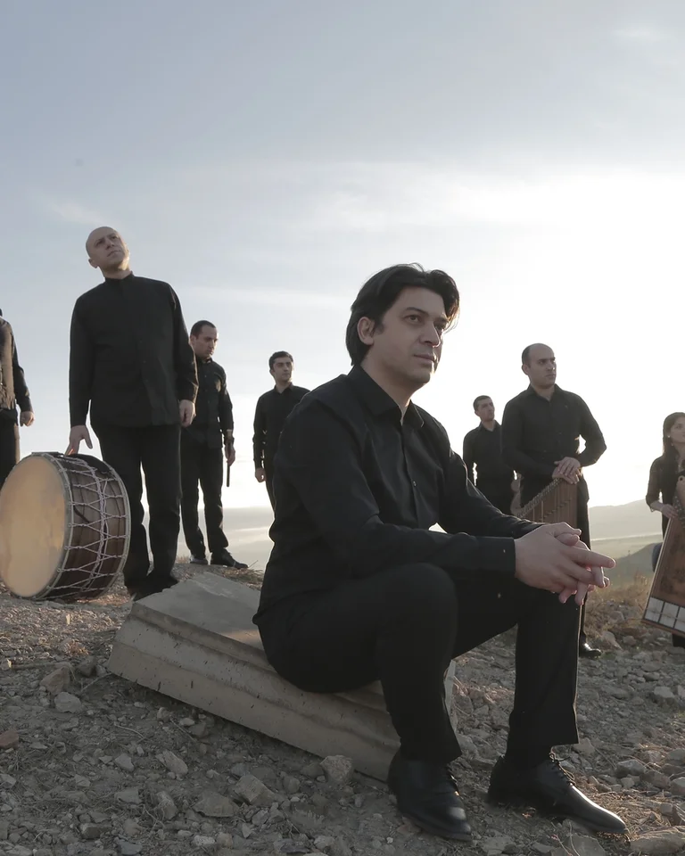 Eine Gruppe von Musikern (Gurdjieff Ensemble) in dunkler Kleidung steht auf einem Hügel bei Sonnenuntergang, einige halten traditionelle Instrumente.