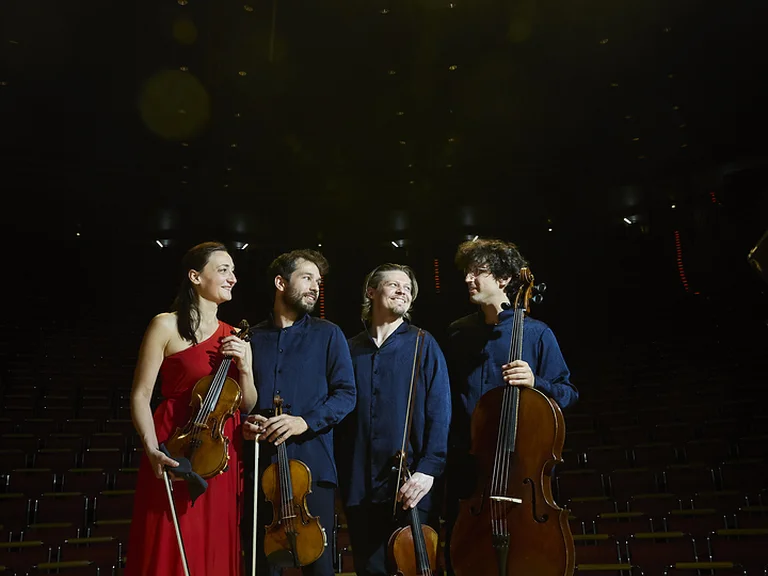 Four musicians (Alinde Quartett) standing on the stage of the Kölner Philharmonie. Two holding cellos, and one with a violin. They are smiling and wearing formal attire.