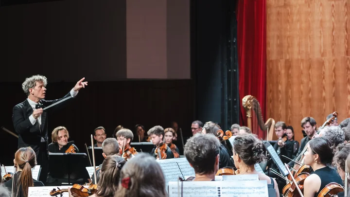Das Mahler Academy Orchestra spielt auf einer Bühne. Der Dirigent (Philipp von Steinaecker) hebt den Arm, um die Musiker zu führen.