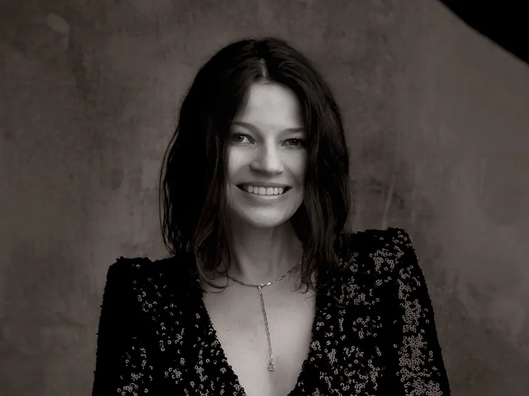 Eine Frau (Dorothee Oberlinger) mit schulterlangem Haar trägt ein glitzerndes schwarzes Kleid und lächelt mit verschränkten Armen vor einem grauen Hintergrund.