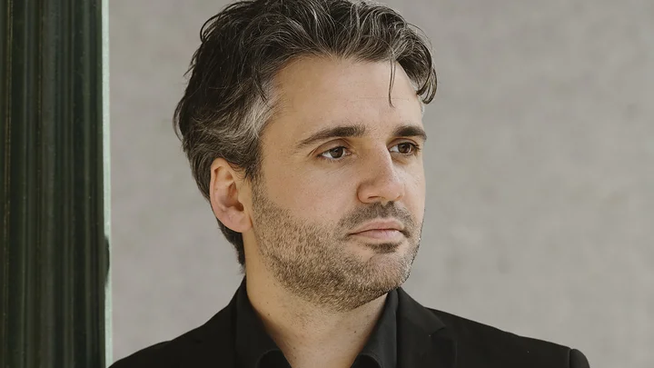 Ein Mann (Reinoud Van Mechelen) mit dunklem, welligem Haar steht in einem schwarzen Anzug vor einer grauen Wand und schaut nach rechts.