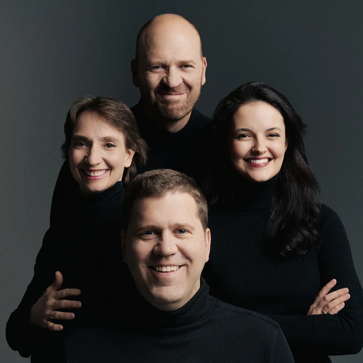 Zwei Männer und zwei Frauen (Cuarteto Casals) in dunkler Kleidung lächeln nebeneinander vor einem grauen Hintergrund.