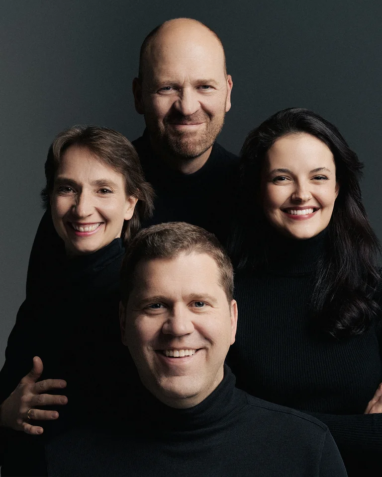 Zwei Männer und zwei Frauen (Cuarteto Casals) in dunkler Kleidung lächeln nebeneinander vor einem grauen Hintergrund.