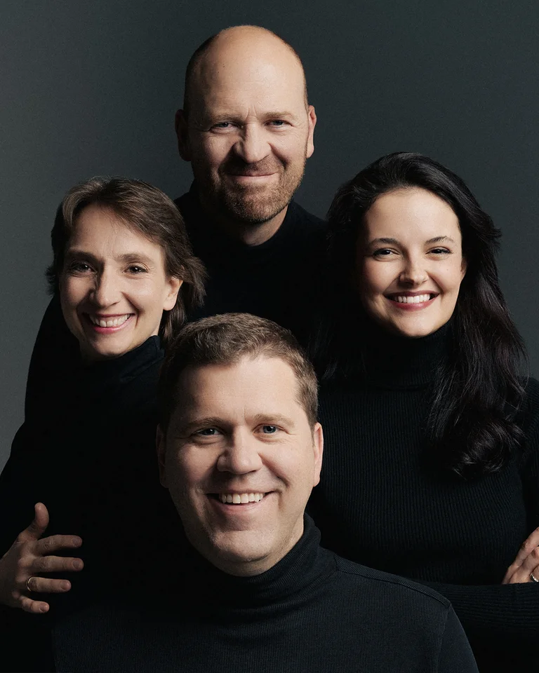 Zwei Männer und zwei Frauen (Cuarteto Casals) in dunkler Kleidung lächeln nebeneinander vor einem grauen Hintergrund.