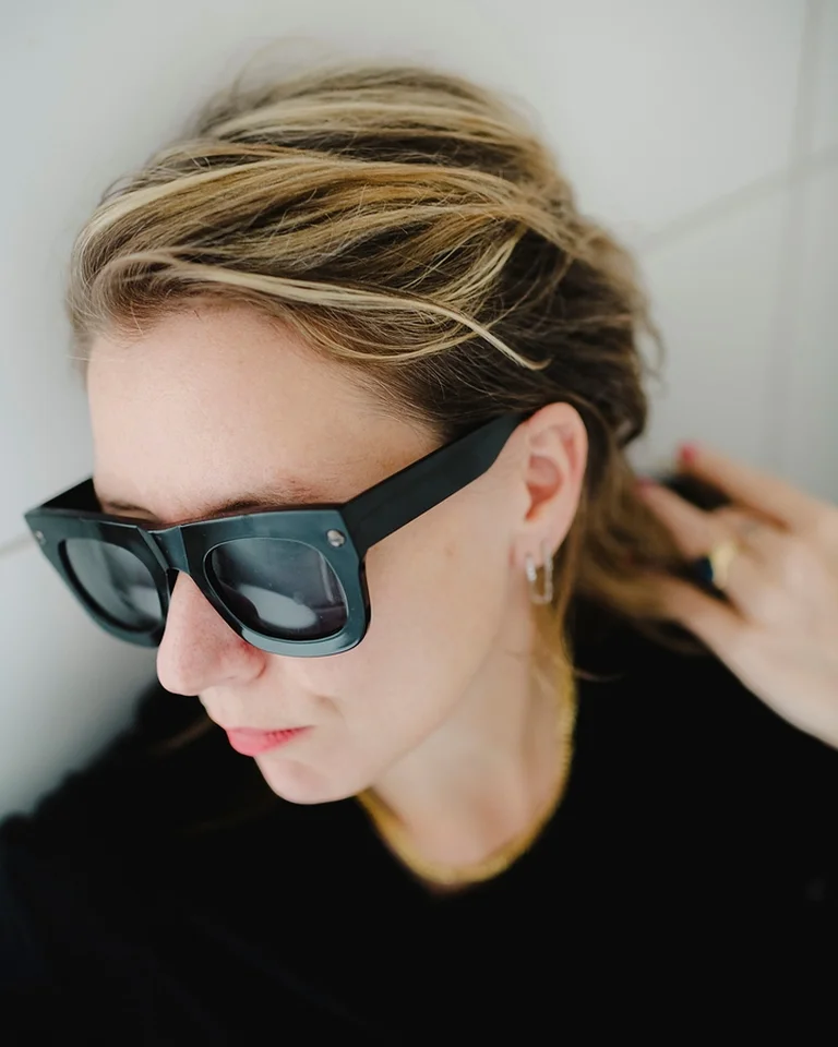 Nahaufnahme einer Frau mit hellbraunem Haar und schwarzen Sonnenbrillen, in Schwarz gekleidet, lehnt sich an eine weiße Wand.
