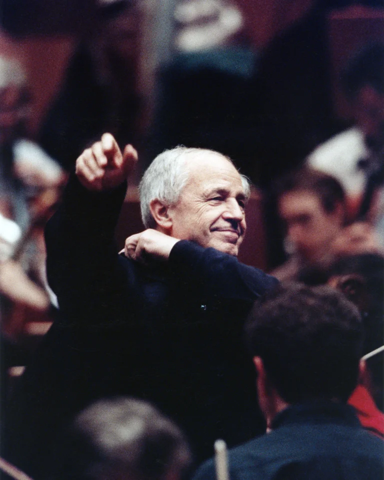 Ein Dirigent mit grauem Haar dirigiert ein Orchester. Seine Gestik zeigt Leidenschaft und Hingabe zur Musik.