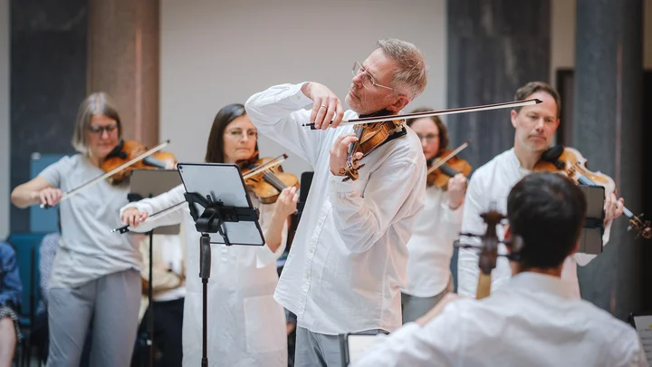 Ein Ensemble von Musikern (Scottish Ensemble) spielt Geige in einem hellen Raum. Sie tragen weiße Kleidung und konzentrieren sich auf ihre Darbietung.