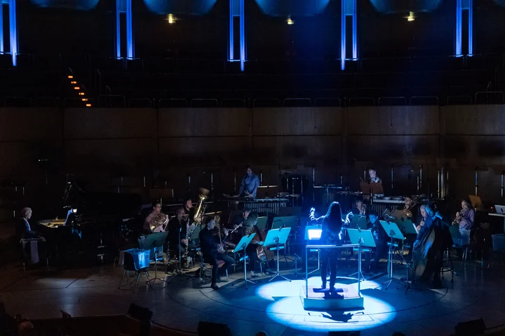 Das SWR Vokalensemble und Ensemble Modern spielen bei unter blauem Licht. Die Dirigentin Xizi Wang steht im Vordergrund, umgeben von den Musiker:innen mit ihren Instrumenten.
