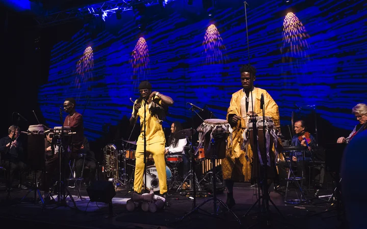 The musicians of the Deggo Band on stage, dressed in yellow, blue, and burgundy, flanked by drums and a blue backdrop. An energetic performance.