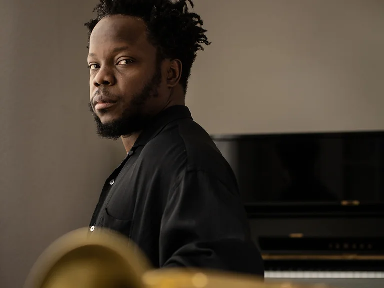 Ein Musiker (Ambrose Akinmusire) sitzt nachdenklich vor einem Klavier, während im Vordergrund ein unscharfes Blasinstrument zu sehen ist.