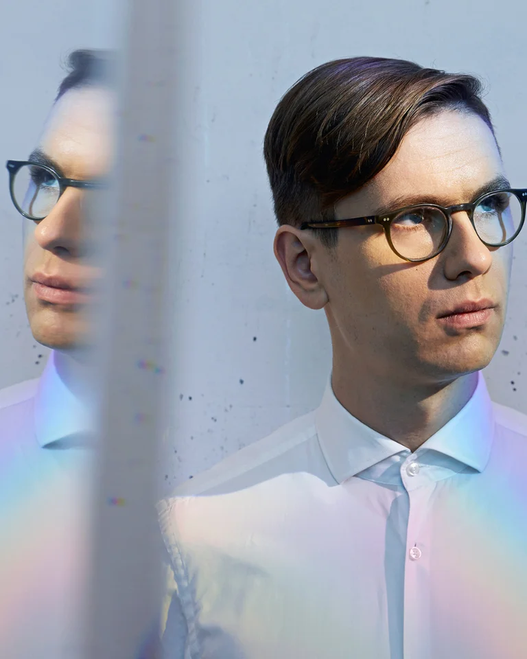 Ein Mann mit Brille und Hemd schaut nach rechts. Ein Prismeneffekt erzeugt Reflexionen auf der Wand und dem Hemd.