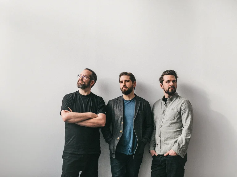Drei Männer (Pablo Held Trio) stehen vor einer weißen Wand. Sie blicken in verschiedene Richtungen und tragen lässige Kleidung in dunklen Tönen.