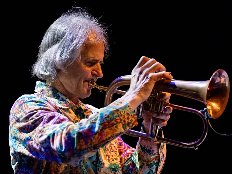 Ein Musiker (Markus Stockhausen) spielt virtuos ein Flügelhorn. Er trägt ein bunt bedrucktes Hemd, der Hintergrund ist dunkel gehalten.