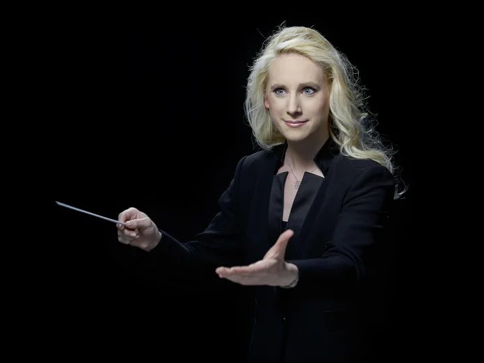 Lena-Lisa Wüstendörfer: Eine Dirigentin mit blonden Haaren in schwarzem Anzug dirigiert leidenschaftlich vor einem dunklen Hintergrund.