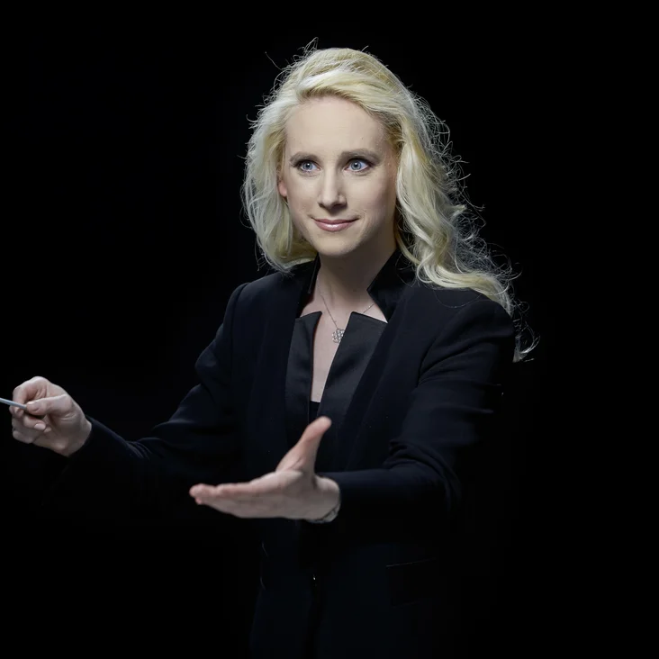 Lena-Lisa Wüstendörfer: Eine Dirigentin mit blonden Haaren in schwarzem Anzug dirigiert leidenschaftlich vor einem dunklen Hintergrund.