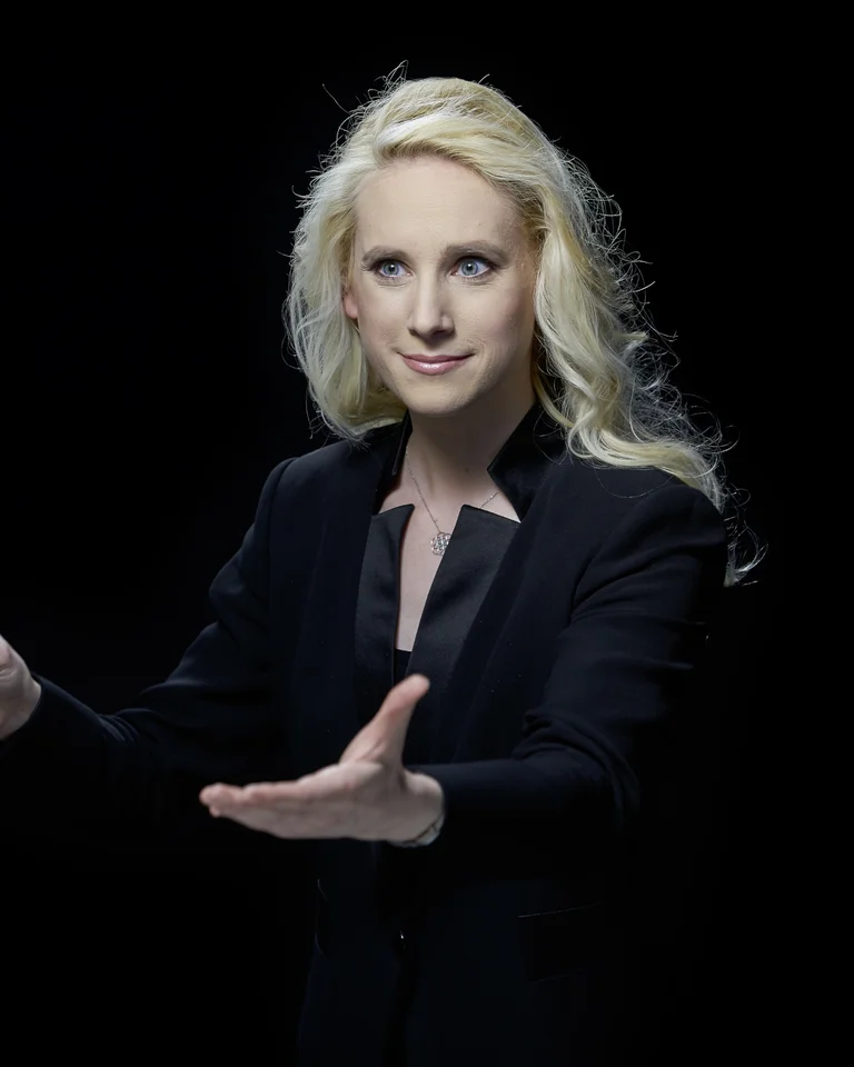 Lena-Lisa Wüstendörfer: Eine Dirigentin mit blonden Haaren in schwarzem Anzug dirigiert leidenschaftlich vor einem dunklen Hintergrund.
