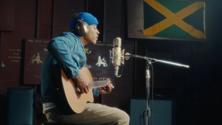 Patrice in a blue cap sings and plays guitar in a studio. A Jamaican flag hangs in the background.