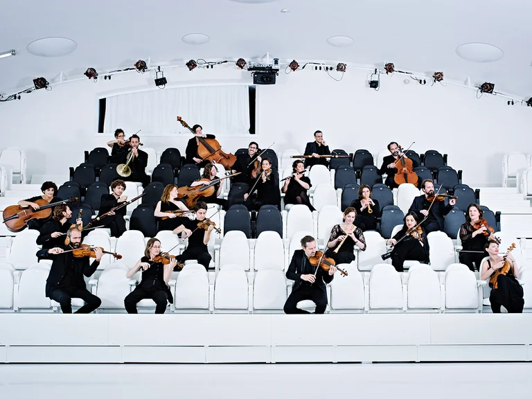 Ein Orchester (Le concert de la Loge) spielt auf weißen Stuhlreihen in einem modernen Saal. Musiker tragen formelle schwarze Kleidung und spielen verschiedene Instrumente.