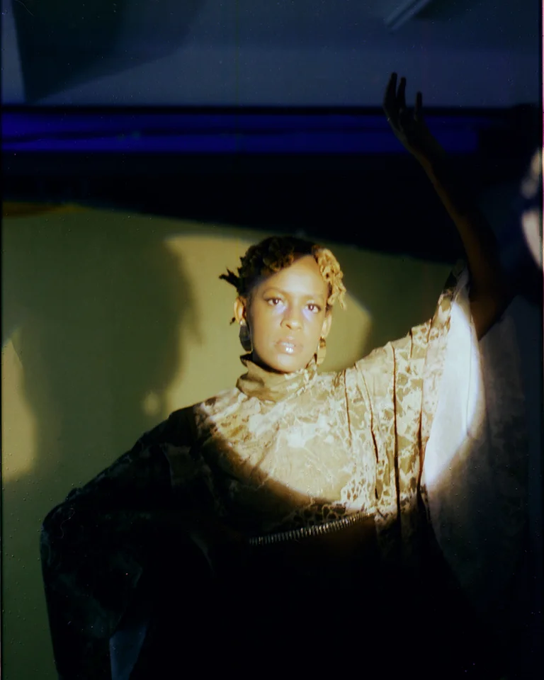 Eine Person in einer dramatischen Pose (Nyokabi Kariũki), beleuchtet von einem kreisförmigen Licht. Der Hintergrund ist in dunklen Tönen gehalten.