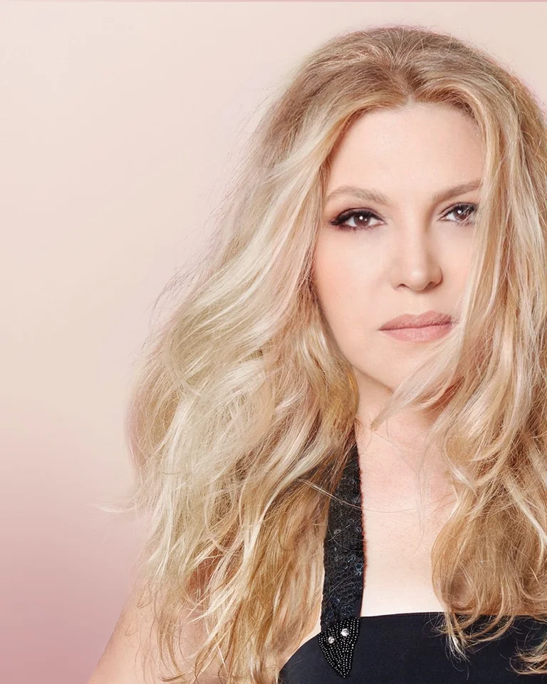 Eine Person mit langen blonden Haaren schaut direkt in die Kamera. Der Hintergrund ist weich und pastellfarben.