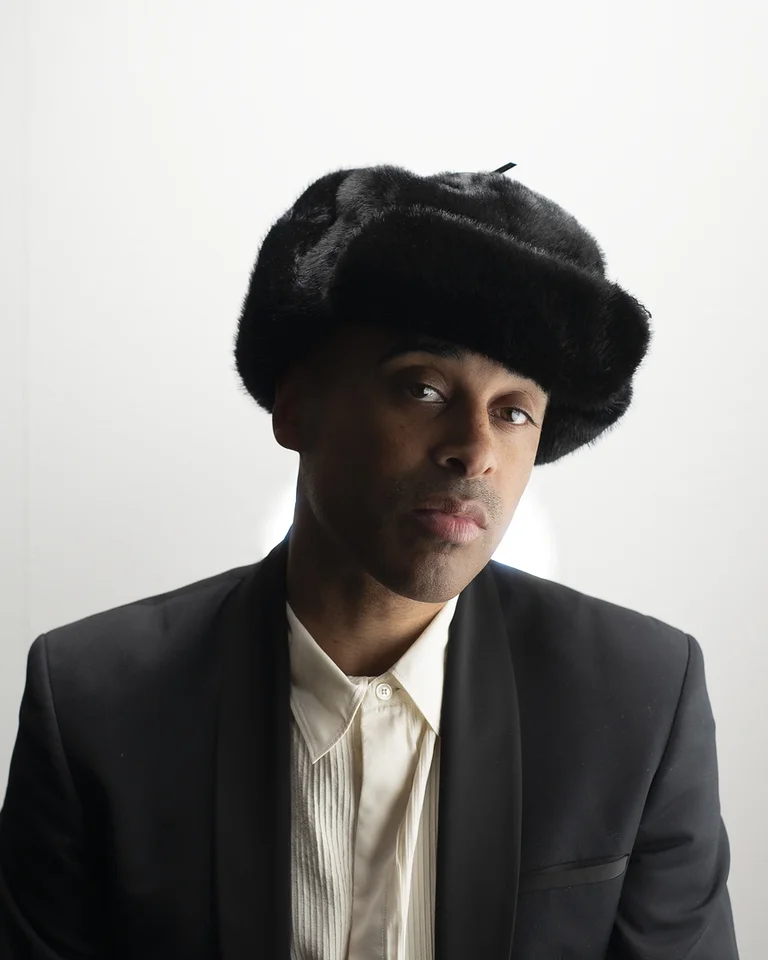 Mann (Patrice) in formellem Anzug und Pelzmütze, vor weißem Hintergrund. Seine Haltung ist ruhig und ausdrucksstark.