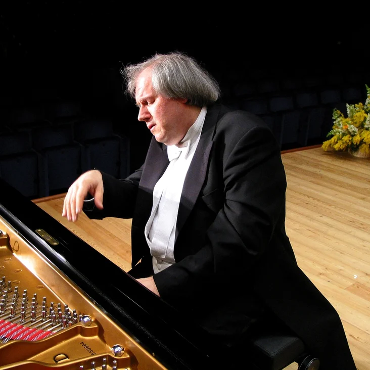 Grigory Sokolov: Ein Pianist mit grauem Haar spielt intensiv auf einem Konzertflügel auf einer Bühne mit Blumenarrangement.