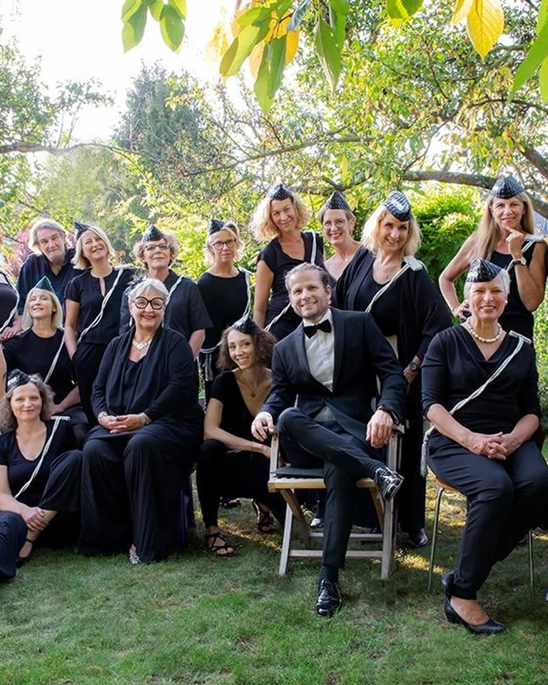 A group of people in black outfits and hats happily posing outdoors under trees for a group photo.