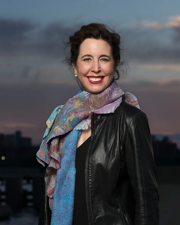 Eine Frau mit lockigem Haar trägt eine lederne Jacke und einen bunten Schal vor einer Stadtlandschaft im Abendlicht.