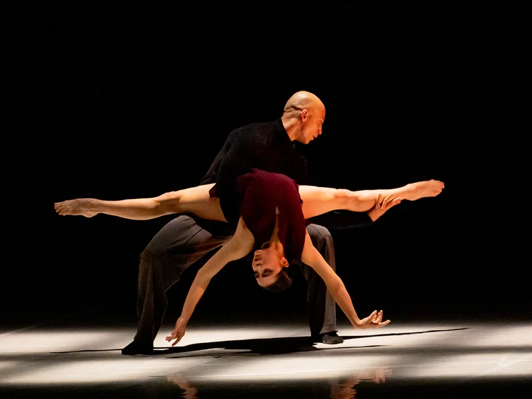 Zwei Tänzer führen eine beeindruckende Hebefigur auf einer Bühne auf. Der Hintergrund ist dunkel, der Boden hell beleuchtet.