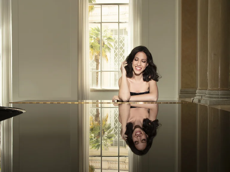 Eine Frau (Beatrice Rana) sitzt an einem Flügel in einem elegant dekorierten Raum mit großen Fenstern. Ihr Spiegelbild ist sichtbar.