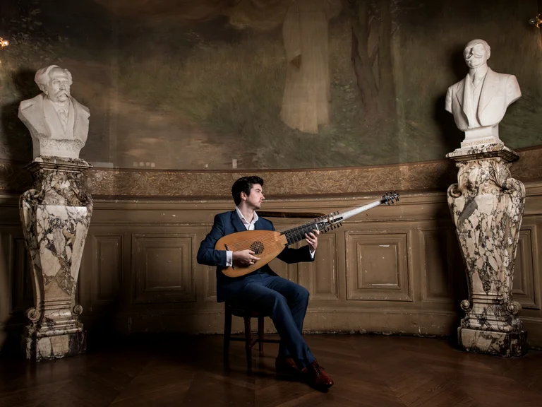 Ein Mann (Thomas Dunford) in einem eleganten Anzug spielt Laute in einem kunstvollen Raum mit Büsten und dekorativen Säulen.