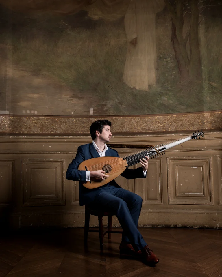 Ein Mann (Thomas Dunford) in einem eleganten Anzug spielt Laute in einem kunstvollen Raum mit Büsten und dekorativen Säulen.