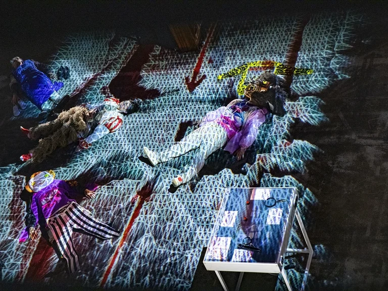 Eine künstlerische Performance mit leuchtenden Projektionen auf einer Bühne. Menschen in kreativ gestalteten Kostümen liegen in verschiedenen Posen.