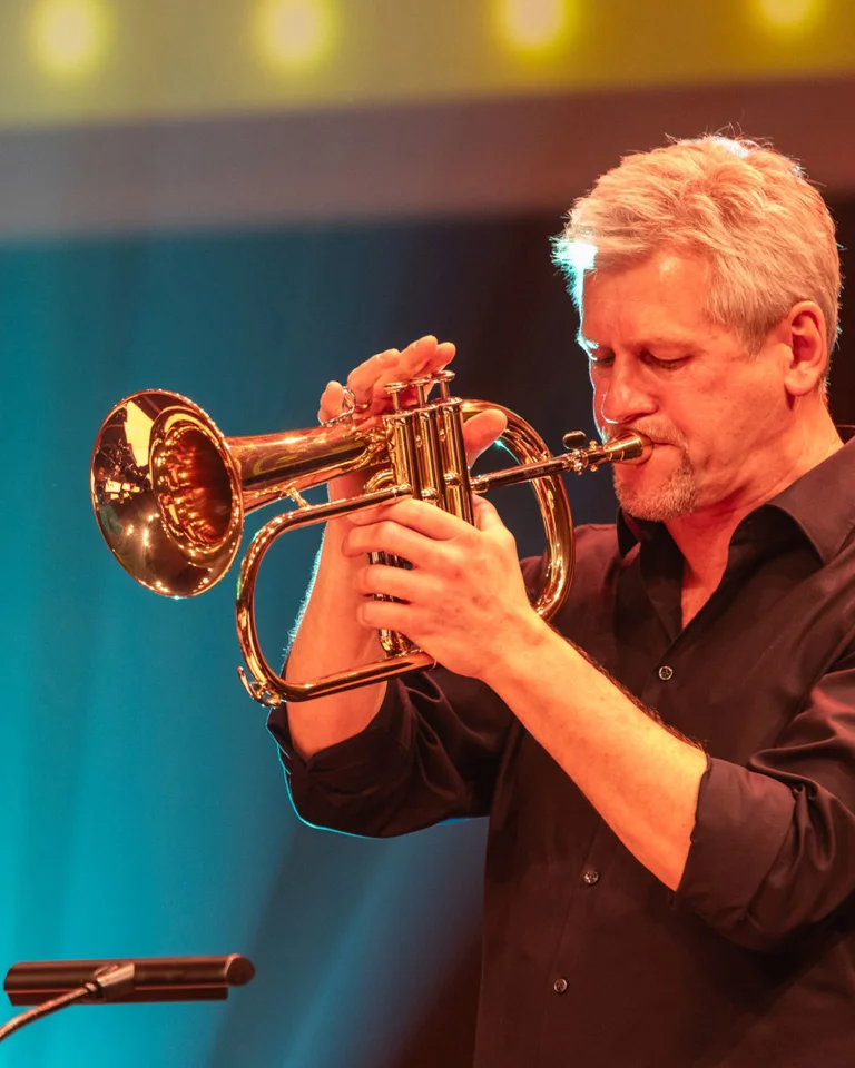 Ein Musiker spielt Trompete auf der Bühne unter leuchtenden Lichtern. Ein Mikrofon steht vor ihm. Stimmung ist festlich.