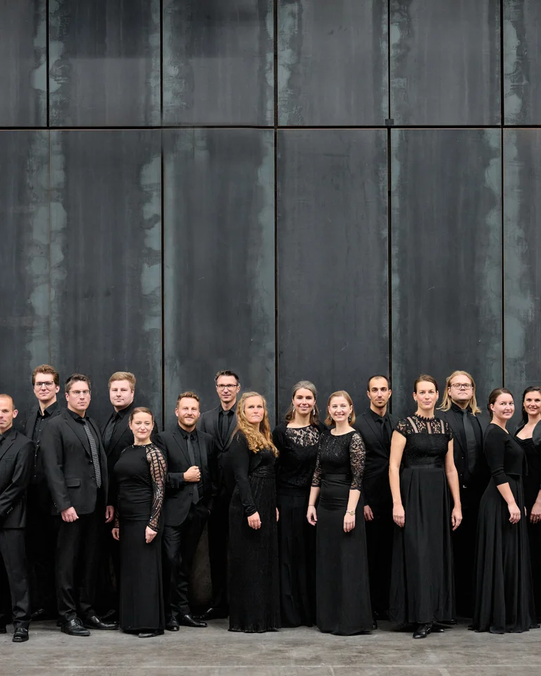 Eine Gruppe von Menschen in schwarzer Kleidung steht vor einer dunklen Wand. Sie posieren nebeneinander und schauen direkt in die Kamera.