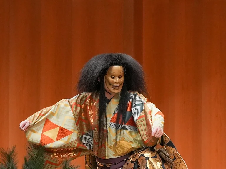 Ein Schauspieler in einem traditionellen Noh-Kostüm (Kokaji) und -Maske führt auf einer hölzernen Bühne eine klassische japanische Aufführung auf.